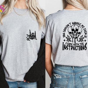 Op de afbeelding: Grijs T-shirt met een zwarte skelet hand die een rock and roll-teken maakt aan de linkerkant en de tekst "You couldn't handle me even if I come with the instructions" met een zwarte schedel afbeelding aan de rechterkant.
