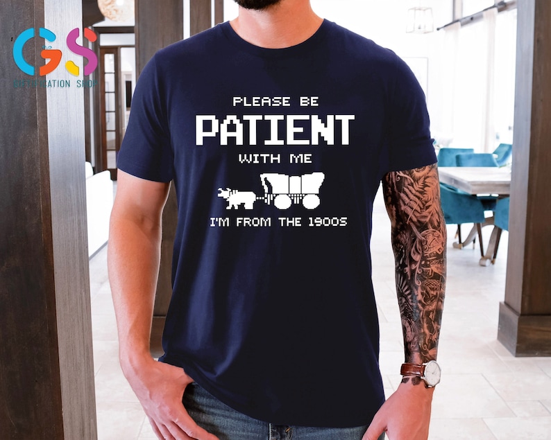 Puede incluir: Una camiseta azul marino con texto blanco que dice "Por favor, ten paciencia conmigo, soy de los a&ntilde;os 1900". El texto est&aacute; en una fuente pixelada. Una imagen blanca de un carro tirado por bueyes est&aacute; debajo del texto.