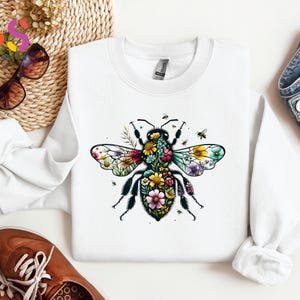 May include: White sweatshirt featuring a colorful floral bee design. The bee's wings are adorned with vibrant flowers in shades of pink, yellow, and purple. The sweatshirt is a casual, comfortable style.