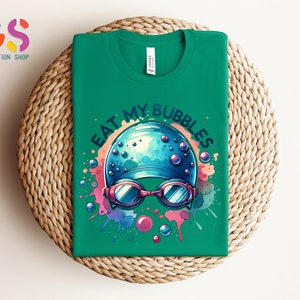 May include: Green t-shirt with a colorful graphic of a swimmer's cap with goggles and the text "Eat My Bubbles".