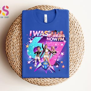 以下が含まれることがあります： グラフィックデザインが施されたロイヤルブルーのTシャツ。「I WAS 67 NOW I'M」の文字と大きな数字の67が特徴です。デザインには漫画のキャラクター、星、青とピンクの配色が含まれています。Tシャツは織り込まれた表面に表示されています。