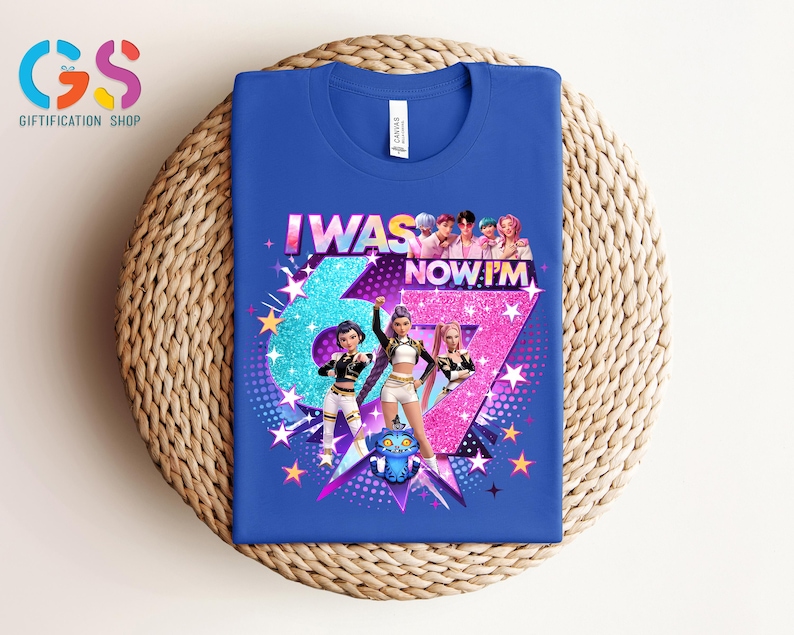 以下が含まれることがあります： グラフィックデザインが施されたロイヤルブルーのTシャツ。「I WAS 67 NOW I'M」の文字と大きな数字の67が特徴です。デザインには漫画のキャラクター、星、青とピンクの配色が含まれています。Tシャツは織り込まれた表面に表示されています。