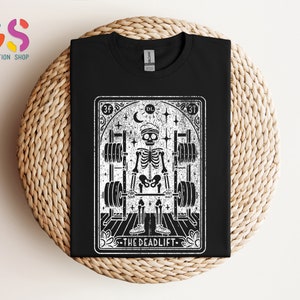 May include: Black and white illustration of a skeleton lifting weights. The illustration is in the style of a tarot card with the text "The Deadlift" at the bottom.