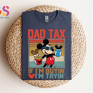 May include: Navy blue t-shirt featuring Mickey Mouse wearing sunglasses and holding food and a drink. The text "DAD TAX" is above the image, and "IF I'M BUYIN' I'M TRYIN'" is below. The shirt is displayed on a woven surface.