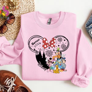 Pode incluir: Um moletom rosa claro com um design da Disney. O design inclui orelhas do Mickey Mouse, um la&ccedil;o vermelho com bolinhas, fogos de artif&iacute;cio e v&aacute;rios personagens da Disney. O nome "Sophia 2026" tamb&eacute;m &eacute; exibido. O moletom &eacute; feito de um material macio.
