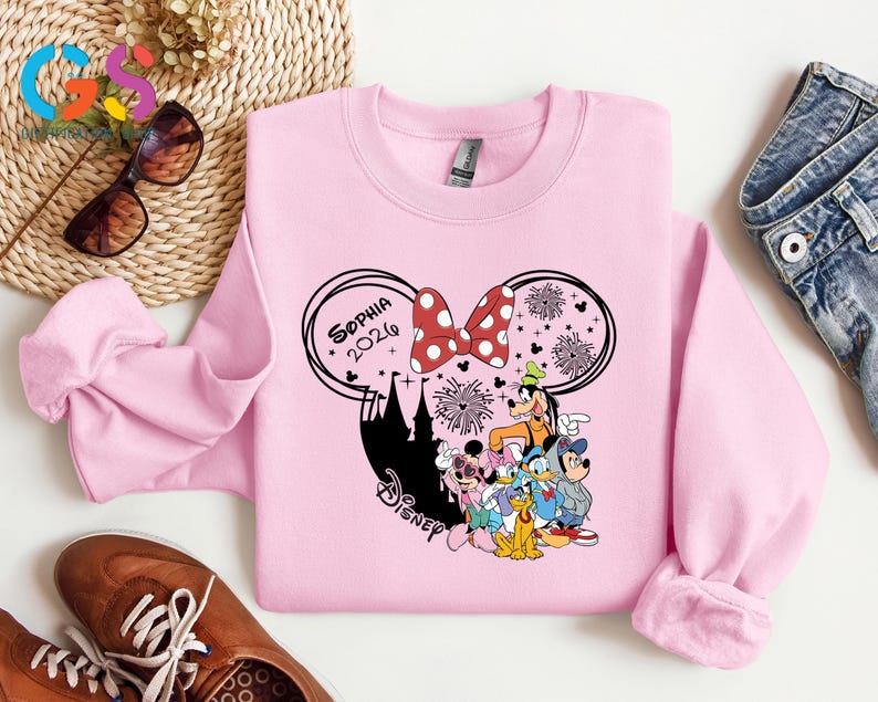 Pode incluir: Um moletom rosa claro com um design da Disney. O design inclui orelhas do Mickey Mouse, um la&ccedil;o vermelho com bolinhas, fogos de artif&iacute;cio e v&aacute;rios personagens da Disney. O nome "Sophia 2026" tamb&eacute;m &eacute; exibido. O moletom &eacute; feito de um material macio.