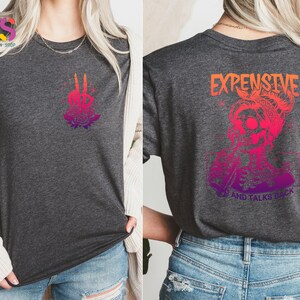 May include: A gray t-shirt with a pink and orange gradient design. The front of the shirt features a skeleton hand with roses. The back of the shirt features a skeleton with a bun and the text "EXPENSIVE AND TALKS BACK".