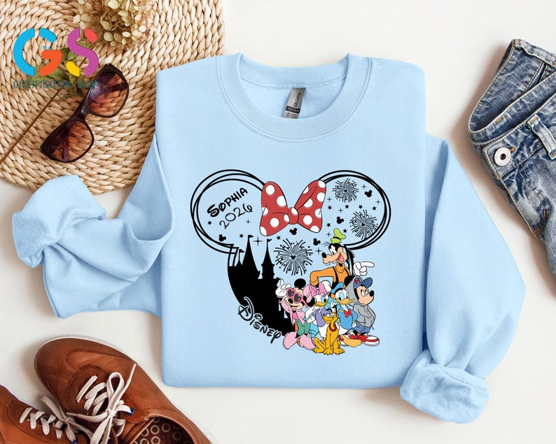 Pode incluir: Camisola azul clara com um design da Disney. O design inclui um la&ccedil;o vermelho e branco com bolinhas, o texto "Sophia 2026", uma silhueta preta de um castelo e personagens da Disney. A camisola &eacute; feita de um material macio.