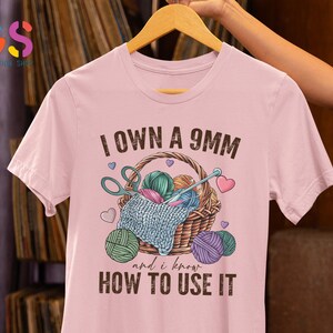 May include: Pink t-shirt with the text "I own a 9mm and I know how to use it". The shirt features a graphic of a basket with yarn, knitting needles, and scissors.