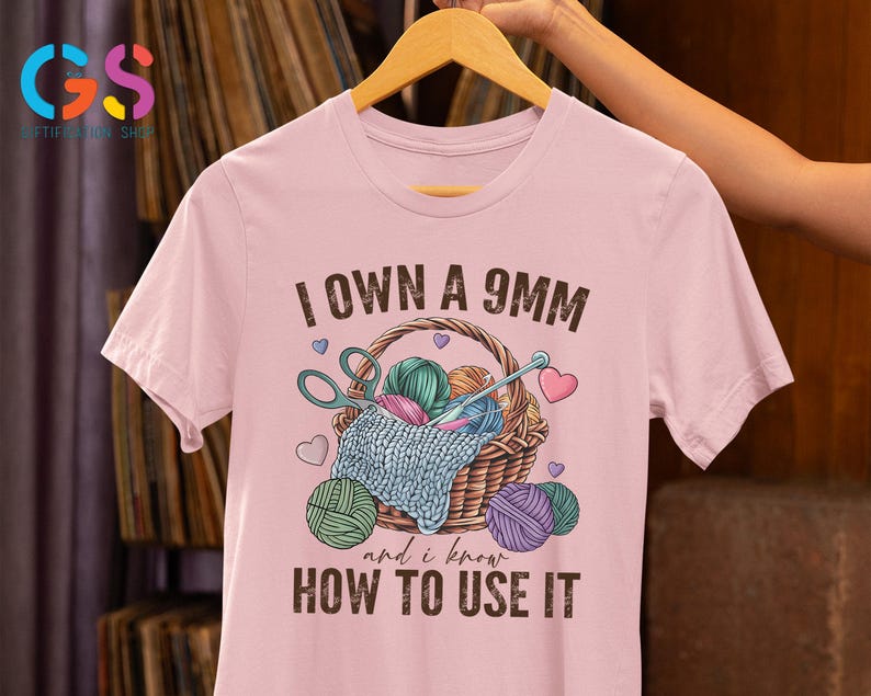 May include: Pink t-shirt with the text "I own a 9mm and I know how to use it". The shirt features a graphic of a basket with yarn, knitting needles, and scissors.