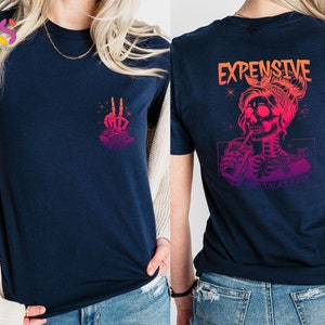 May include: A navy blue t-shirt with a pink and orange gradient design. The front of the shirt features a skeleton hand making a peace sign with a rose. The back of the shirt features a skeleton woman with a drink and the text "Expensive and Talks Back".