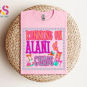 Puede incluir: Camiseta rosa claro con la frase "RUNNING ON ALANI & CHAOS" en un dise&ntilde;o colorido. El dise&ntilde;o incluye ilustraciones de un loro, un c&oacute;ctel, un flamenco y una rodaja de sand&iacute;a. La camiseta est&aacute; doblada sobre una superficie tejida.