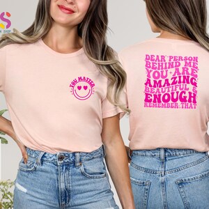 May include: Two pink t-shirts with a positive message. The front shirt says "You Matter" with a smiley face and hearts. The back shirt says "Dear Person Behind Me You Are Amazing Beautiful & Enough Remember That".