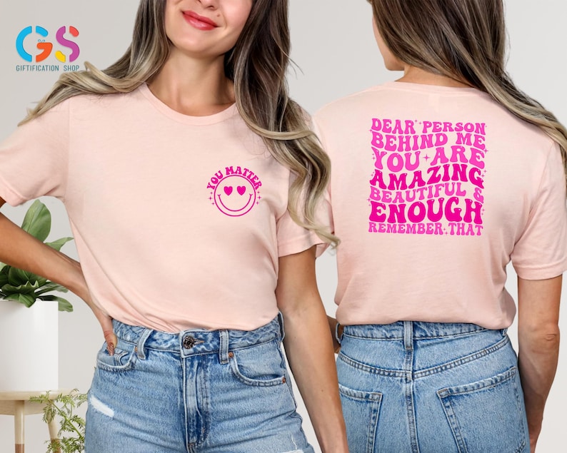 May include: Two pink t-shirts with a positive message. The front shirt says "You Matter" with a smiley face and hearts. The back shirt says "Dear Person Behind Me You Are Amazing Beautiful & Enough Remember That".