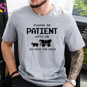 Puede incluir: Camiseta gris con un dise&ntilde;o pixelado negro que dice "Por favor, ten paciencia conmigo, soy de los a&ntilde;os 1900". El dise&ntilde;o incluye una imagen pixelada de un carro tirado por un buey.