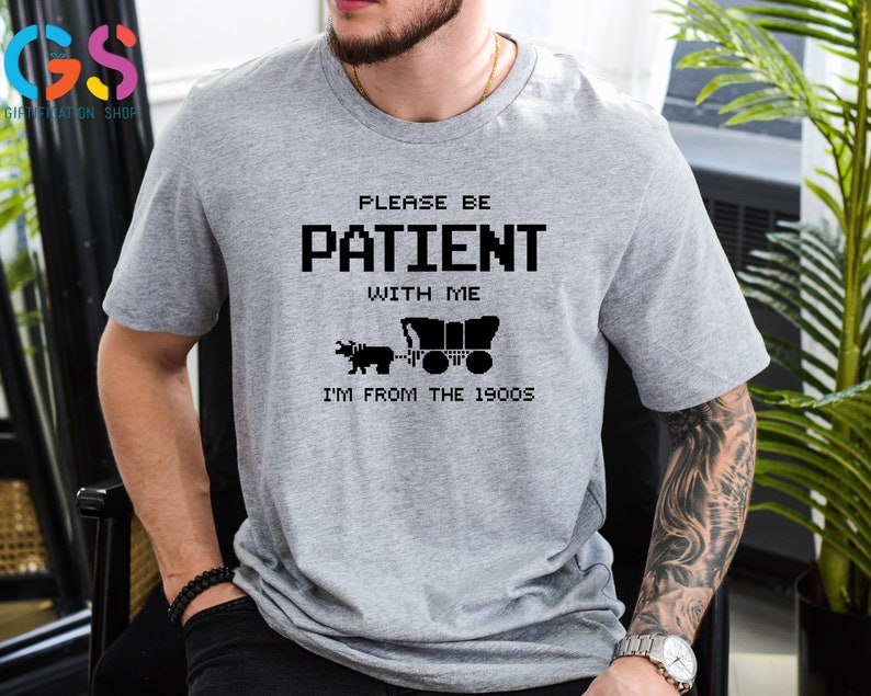 Puede incluir: Camiseta gris con un dise&ntilde;o pixelado negro que dice "Por favor, ten paciencia conmigo, soy de los a&ntilde;os 1900". El dise&ntilde;o incluye una imagen pixelada de un carro tirado por un buey.