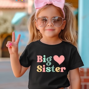 May include: A young girl wearing a black t-shirt with the text "Big Sister" in colorful letters and a pink heart. She is wearing a pink headband and sunglasses.