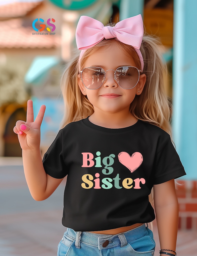 May include: A young girl wearing a black t-shirt with the text "Big Sister" in colorful letters and a pink heart. She is wearing a pink headband and sunglasses.