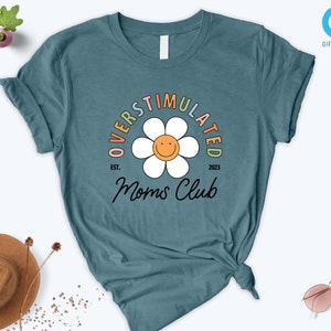 May include: Teal blue t-shirt with a white daisy graphic and the text "Overstimulated Moms Club est. 2023".