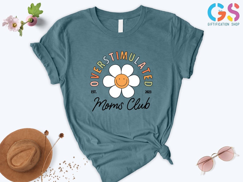 May include: Teal blue t-shirt with a white daisy graphic and the text "Overstimulated Moms Club est. 2023".