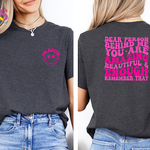 May include: A dark gray t-shirt with pink text that reads "Dear person behind me you are amazing beautiful & enough remember that" on the back and a pink smiley face with hearts for eyes that reads "You matter" on the front.