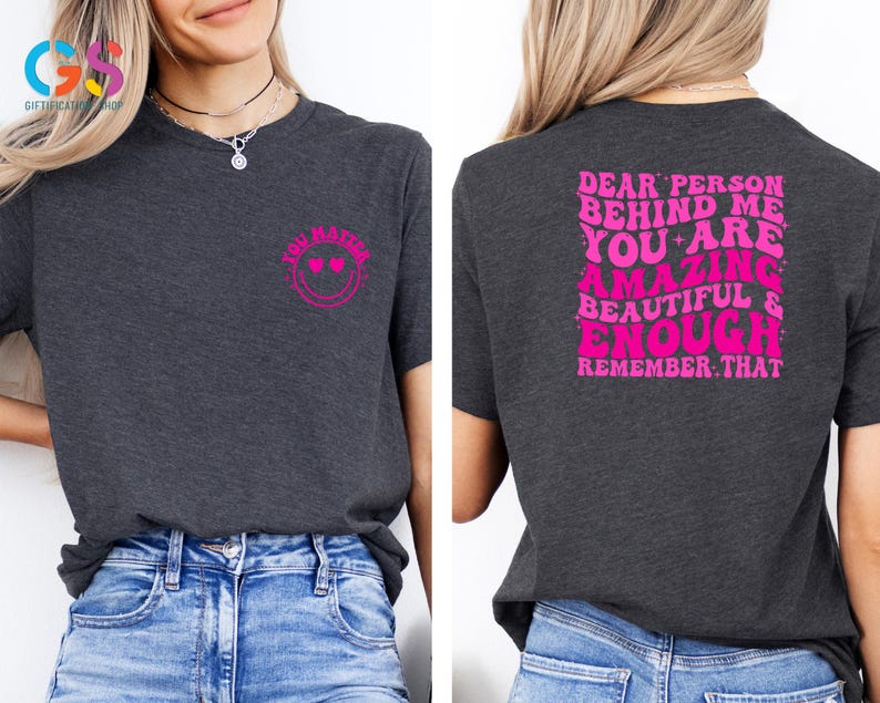 May include: A dark gray t-shirt with pink text that reads "Dear person behind me you are amazing beautiful & enough remember that" on the back and a pink smiley face with hearts for eyes that reads "You matter" on the front.
