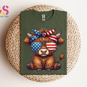 Puede incluir: Camiseta verde oliva con una vaca de las Tierras Altas de dibujos animados que lleva gafas de sol y una bandana, ambas decoradas con la bandera americana. La vaca tiene pelaje marr&oacute;n y un piercing en la nariz. La camiseta se muestra sobre una superficie tejida.