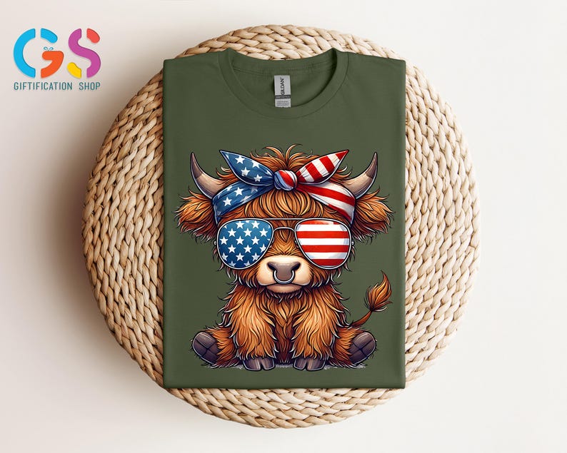 Puede incluir: Camiseta verde oliva con una vaca de las Tierras Altas de dibujos animados que lleva gafas de sol y una bandana, ambas decoradas con la bandera americana. La vaca tiene pelaje marr&oacute;n y un piercing en la nariz. La camiseta se muestra sobre una superficie tejida.