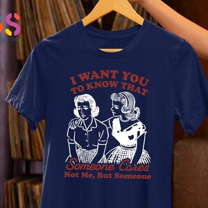 Op de afbeelding: Een marineblauw t-shirt met een witte afbeelding van twee vrouwen. De tekst op het shirt luidt "Ik wil dat je weet dat iemand geeft, niet ik, maar iemand."