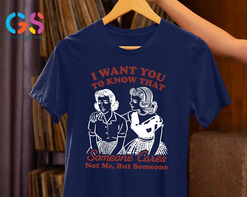 Op de afbeelding: Een marineblauw t-shirt met een witte afbeelding van twee vrouwen. De tekst op het shirt luidt "Ik wil dat je weet dat iemand geeft, niet ik, maar iemand."