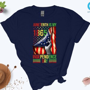 Puede incluir: Camiseta azul marino con un dise&ntilde;o gr&aacute;fico que celebra el Juneteenth. El dise&ntilde;o incluye el texto "Juneteenth is my 1865 Independence Day" con una bandera estadounidense y una mano sosteniendo la bandera. El texto es en rojo, amarillo, verde y negro.
