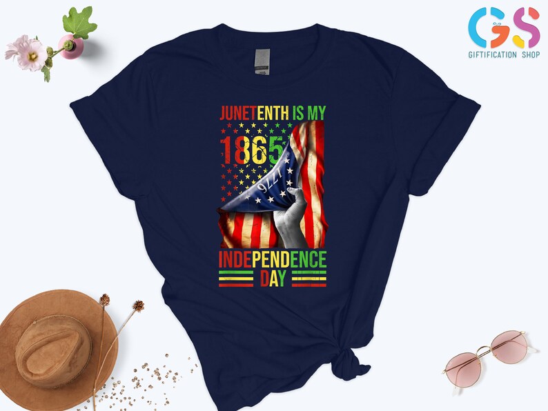 Puede incluir: Camiseta azul marino con un dise&ntilde;o gr&aacute;fico que celebra el Juneteenth. El dise&ntilde;o incluye el texto "Juneteenth is my 1865 Independence Day" con una bandera estadounidense y una mano sosteniendo la bandera. El texto es en rojo, amarillo, verde y negro.