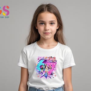 以下が含まれることがあります： 「I WAS 6 NOW I'M 7」の文字が入ったカラフルなグラフィックデザインの白いTシャツ。2人の女の子と星のイラストが幾何学模様の背景に配置されています。Tシャツを着ているのは若い女の子です。