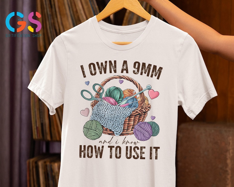May include: White t-shirt with a brown basket filled with yarn balls and a pair of scissors. The text "I own a 9mm and I know how to use it" is printed on the shirt.