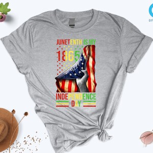 Puede incluir: Una camiseta gris con un dise&ntilde;o gr&aacute;fico del Juneteenth. El dise&ntilde;o incluye el texto "Juneteenth is my 1865 Independence Day" con una bandera estadounidense y una silueta. Una prenda de vestir informal.