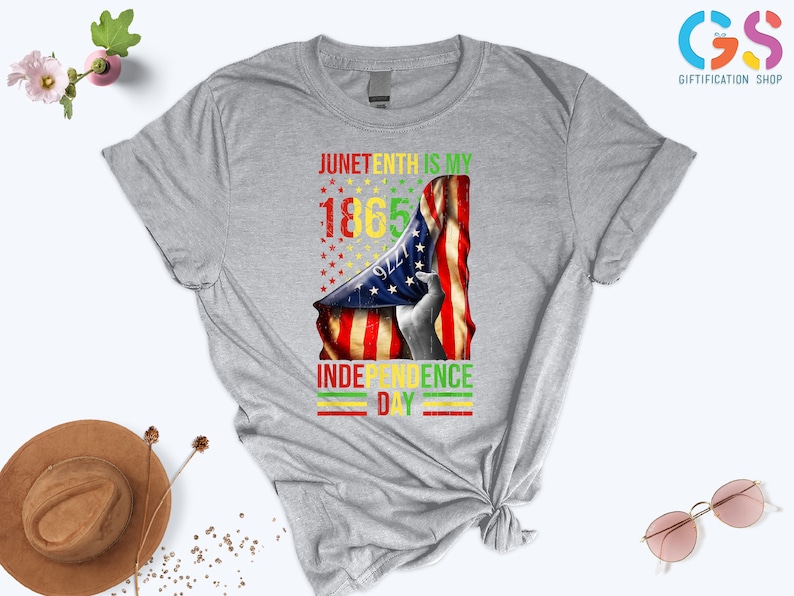 Puede incluir: Una camiseta gris con un dise&ntilde;o gr&aacute;fico del Juneteenth. El dise&ntilde;o incluye el texto "Juneteenth is my 1865 Independence Day" con una bandera estadounidense y una silueta. Una prenda de vestir informal.