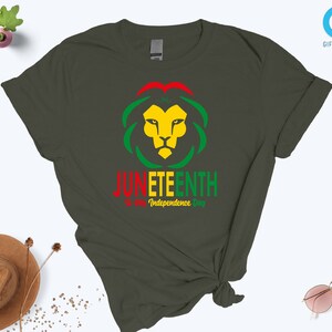 Pode incluir: T-shirt verde escuro com um desenho de cabe&ccedil;a de le&atilde;o em amarelo, vermelho e verde, com as palavras "Juneteenth" e "Independence Day" impressas abaixo. Uma pe&ccedil;a de roupa casual.
