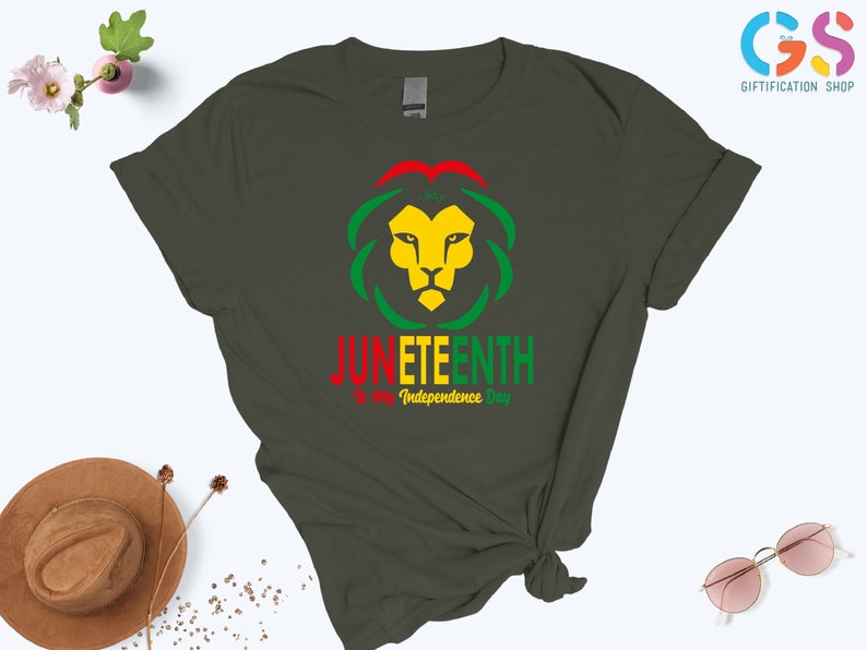 Pode incluir: T-shirt verde escuro com um desenho de cabe&ccedil;a de le&atilde;o em amarelo, vermelho e verde, com as palavras "Juneteenth" e "Independence Day" impressas abaixo. Uma pe&ccedil;a de roupa casual.