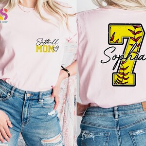 May include: Pink softball mom t-shirt with a gold glitter design that says "Softball Mom" with a heart. The back of the shirt has a yellow softball with red stitching and the number 7 with the name "Sophia" written in black cursive.