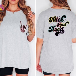 Op de afbeelding: Grijs T-shirt met een zwarte skelethand-afbeelding aan de voorkant en de tekst "Thick And Tired" in een retro-lettertype aan de achterkant.