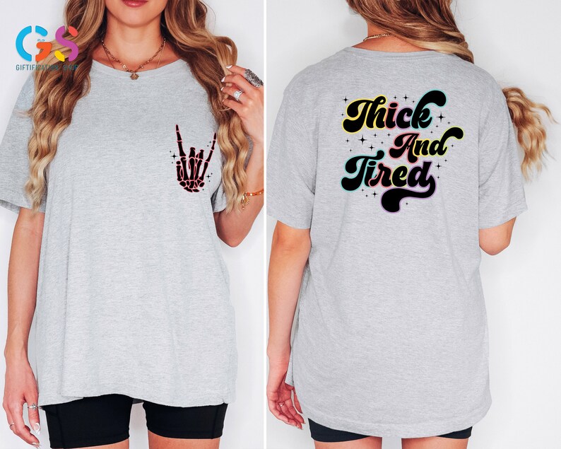 Op de afbeelding: Grijs T-shirt met een zwarte skelethand-afbeelding aan de voorkant en de tekst "Thick And Tired" in een retro-lettertype aan de achterkant.