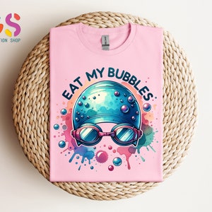 May include: Pink t-shirt with a colorful graphic of a swimmer's cap and goggles with the text "Eat My Bubbles".