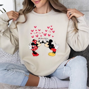May include: Beige sweatshirt featuring a graphic of Mickey and Minnie Mouse kissing, with red hearts floating above them. Mickey Mouse is wearing red shorts and yellow shoes, while Minnie Mouse is in a red polka dot dress and bow. The word "Minnie" is above the castle.