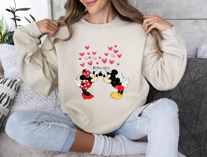May include: Beige sweatshirt featuring a graphic of Mickey and Minnie Mouse kissing, with red hearts floating above them. Mickey Mouse is wearing red shorts and yellow shoes, while Minnie Mouse is in a red polka dot dress and bow. The word "Minnie" is above the castle.