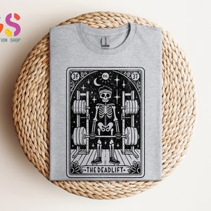 May include: Grey t-shirt featuring a black and white illustration of a skeleton lifting weights. The illustration is within a card-like frame with the text "The Deadlift" at the bottom.