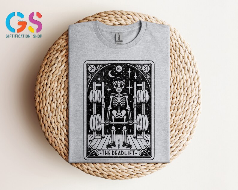 May include: Grey t-shirt featuring a black and white illustration of a skeleton lifting weights. The illustration is within a card-like frame with the text "The Deadlift" at the bottom.