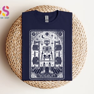 May include: A navy blue t-shirt featuring a white graphic of a skeleton lifting weights. The graphic is in the style of a tarot card and says "The Deadlift" at the bottom.