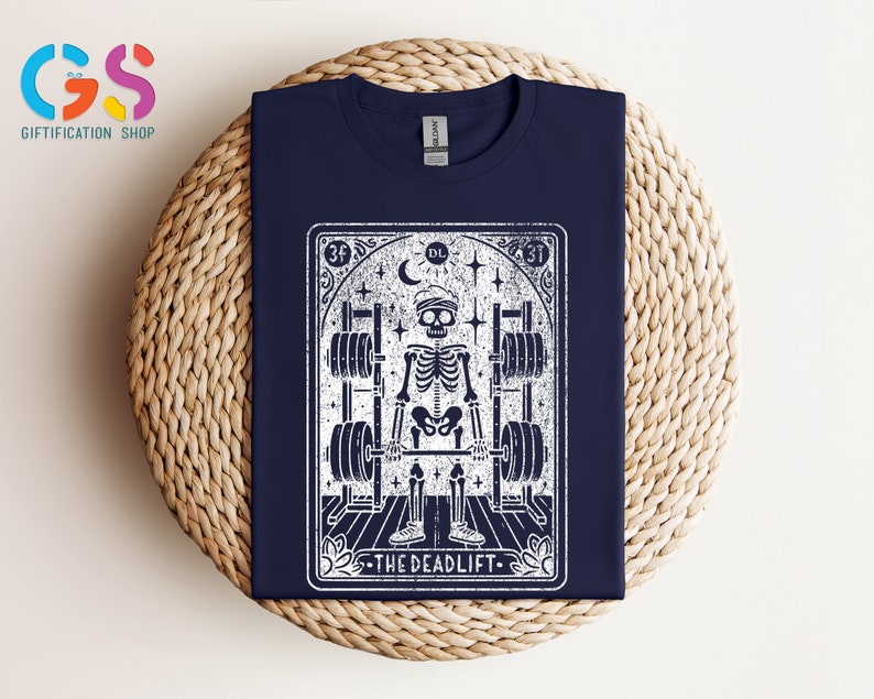 May include: A navy blue t-shirt featuring a white graphic of a skeleton lifting weights. The graphic is in the style of a tarot card and says "The Deadlift" at the bottom.