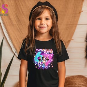 以下が含まれることがあります： 「I WAS... NOW I'M」の文字とカラフルなグラフィックデザインが施された黒いTシャツを着た少女。デザインには星と大きな数字の6が含まれています。彼女は黒いニット帽をかぶっています。