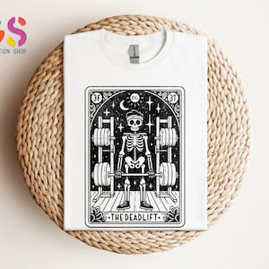 May include: White t-shirt featuring a black and white illustration of a skeleton lifting weights. The illustration is framed in a black border with the text "The Deadlift" at the bottom.